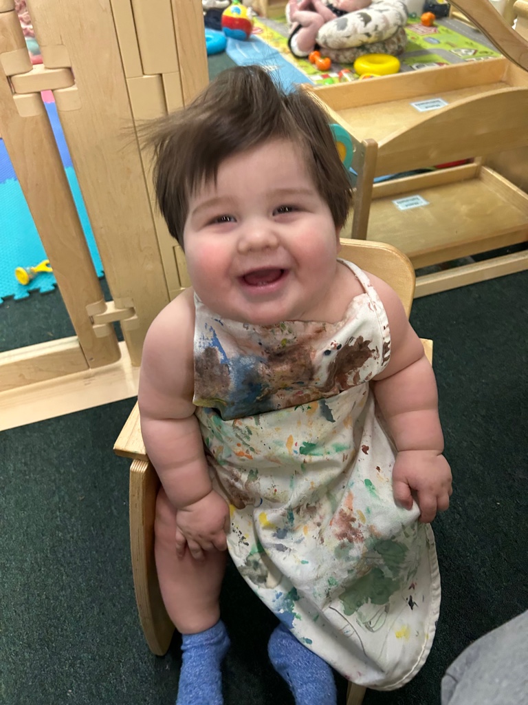 Baby grinning with paint covered art apron